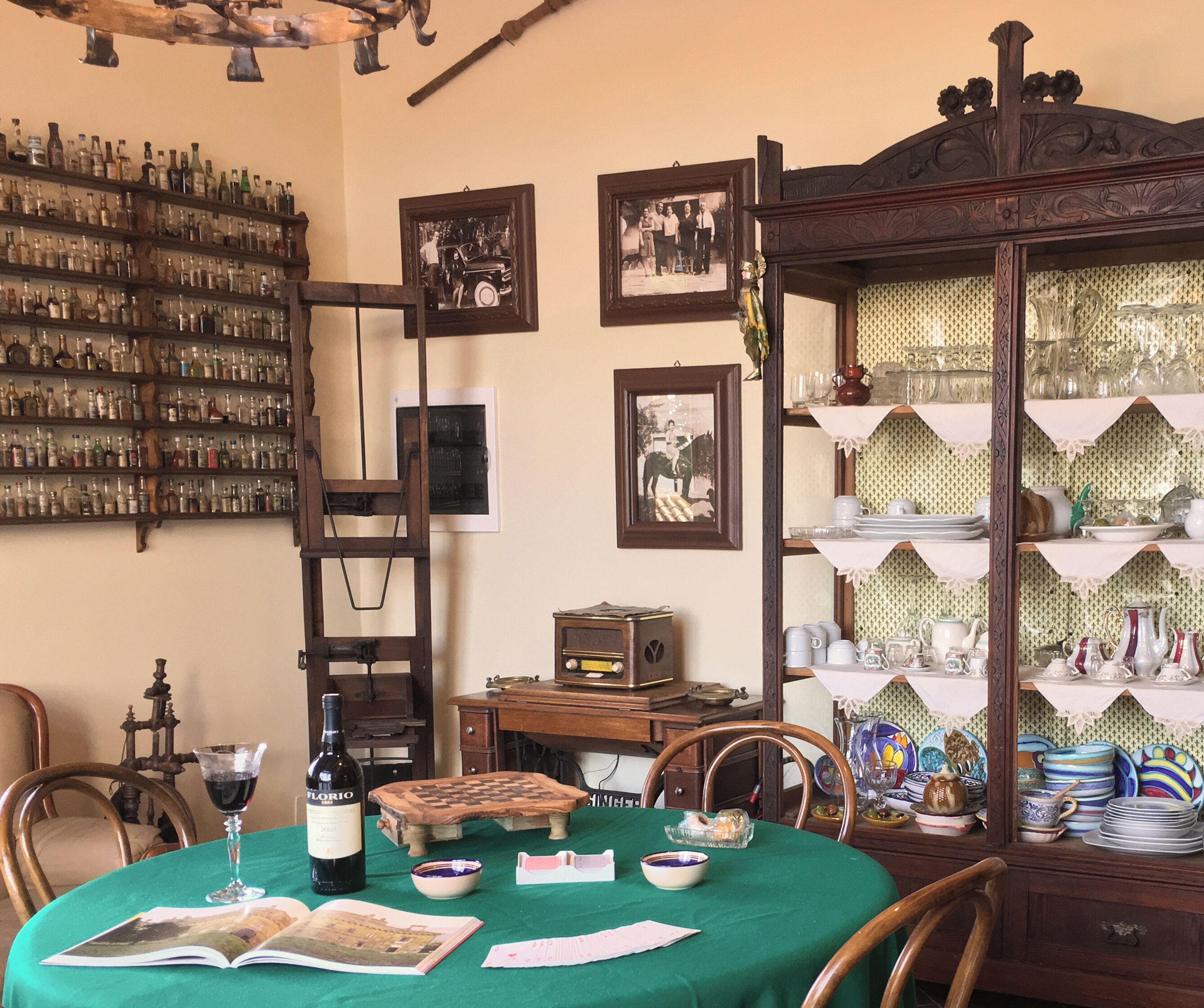 A dining area with a table set for two, a wine bottle, and a book. There are chairs, a wooden cabinet with glassware, and a wall with framed pictures.