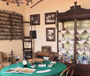 A dining area with a table set for two, a wine bottle, and a book. There are chairs, a wooden cabinet with glassware, and a wall with framed pictures. - Candalù - Villa Giardinata (Valderice)