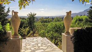 Un patio de piedra con dos grandes jarrones sobre pedestales, rodeado de follaje y vistas a colinas distantes.