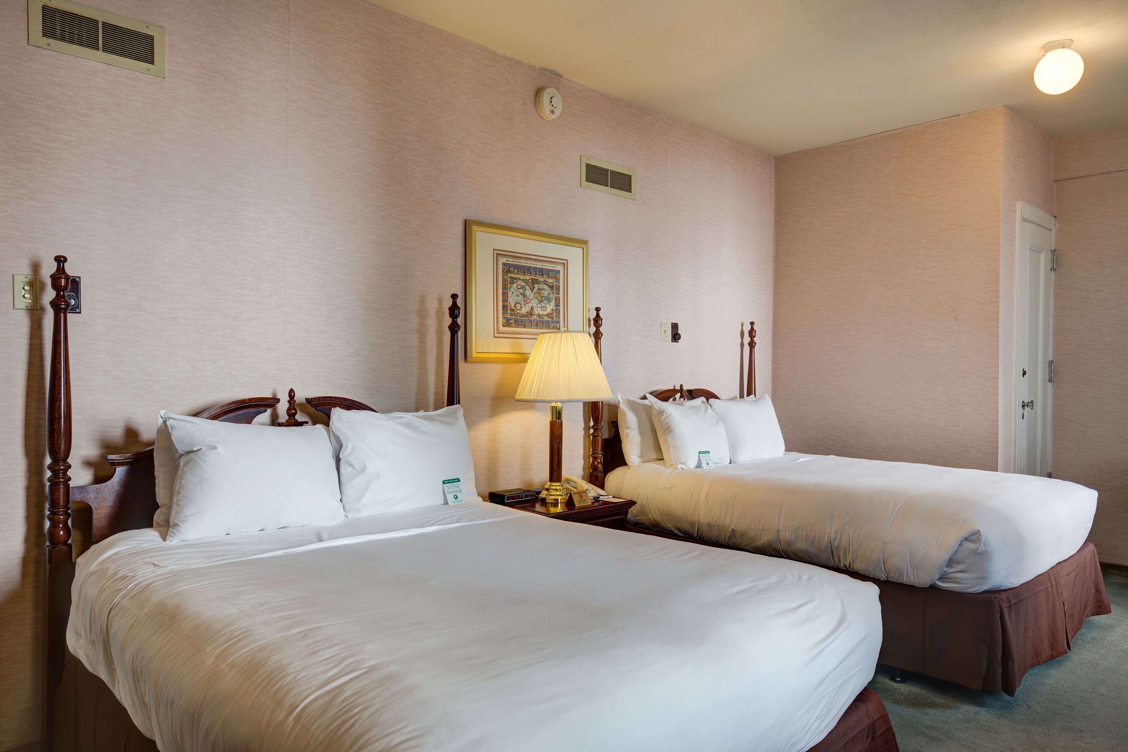 A hotel room with two beds, a bedside table with a lamp, a clock, and a framed picture on the wall.