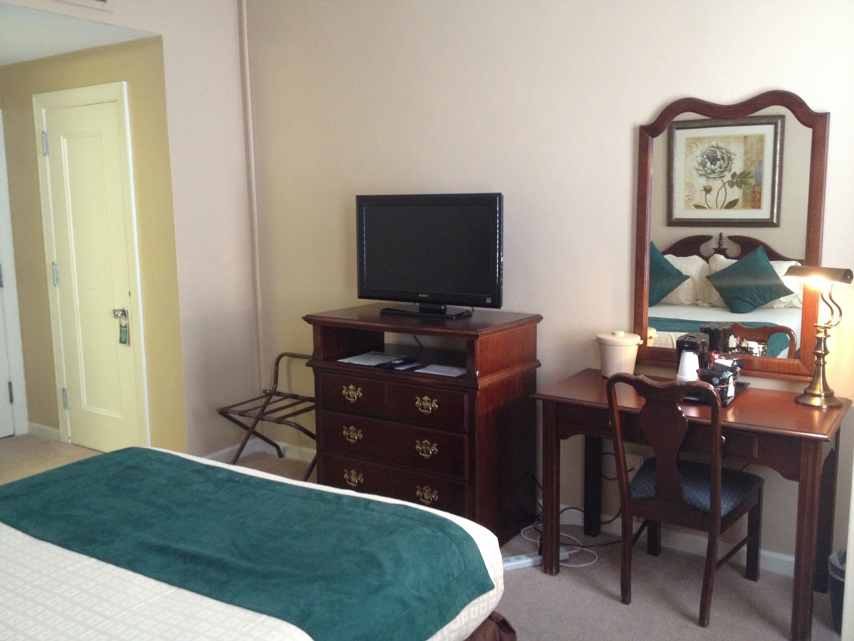 Habitación de hotel con una cama, un escritorio con silla, televisor, cómoda y mesita de noche con lámpara.