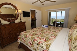 A bedroom with a large bed, a dresser, a mirror, a ceiling fan, and a view of the ocean through a sliding door. - Beach Castle Resort by RVA (Longboat Key)