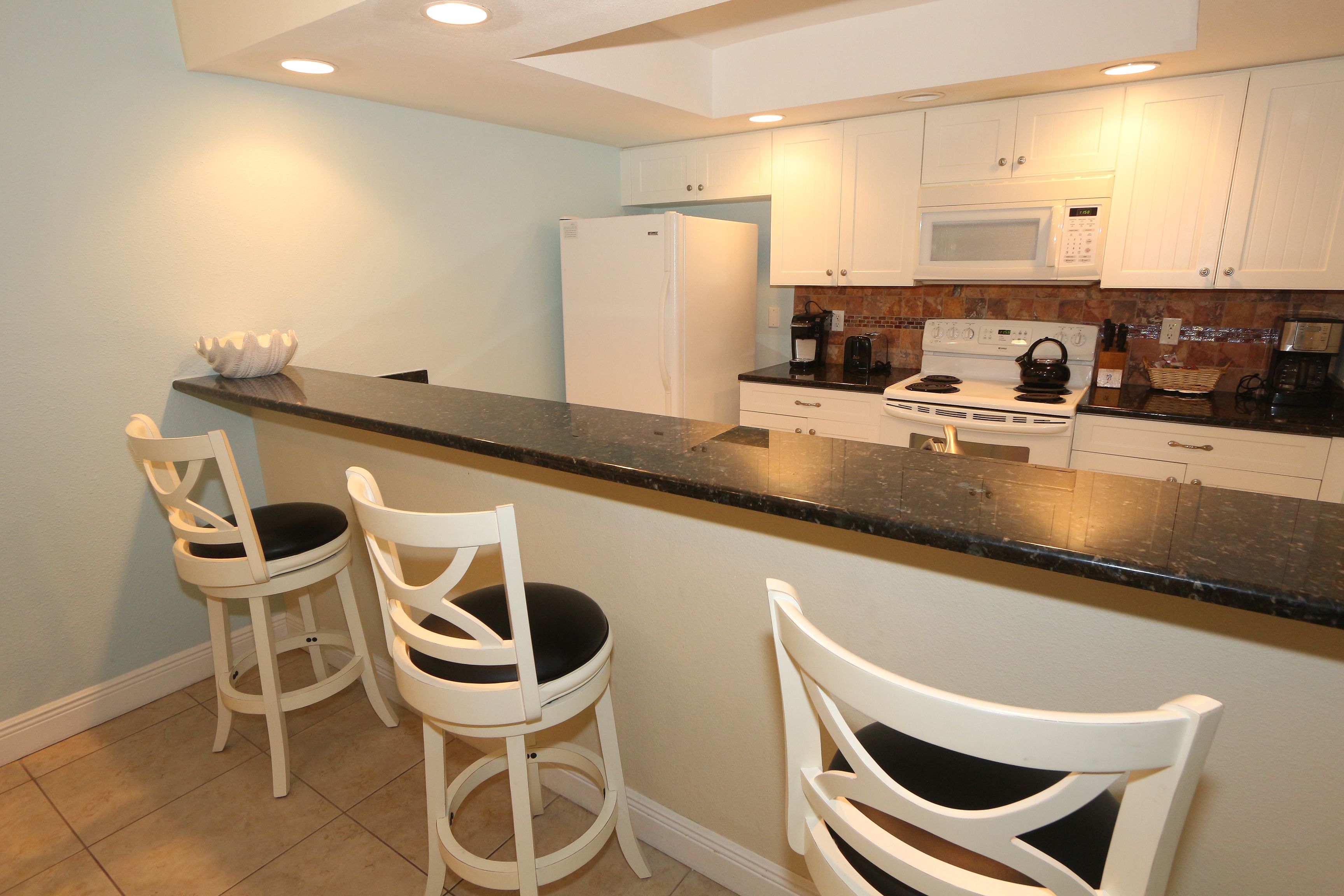 A kitchen with white cabinets, a black countertop, and two white bar stools.