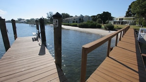 A wooden pier leading to a dock with a 'No Trespassing' sign, a bench, and a clear view of a residential area with houses and a beach.