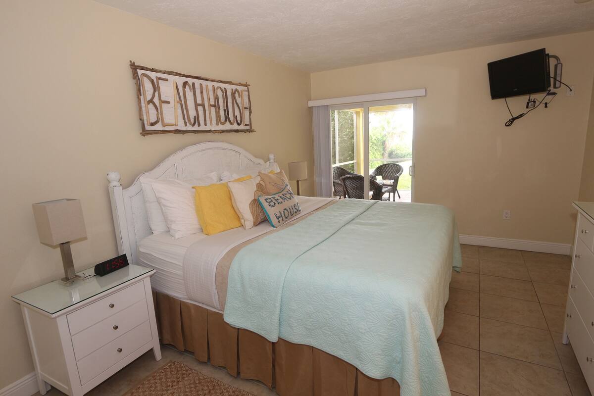 A bedroom with a bed, a nightstand, a TV mounted on the wall, and a sliding glass door leading to a balcony.