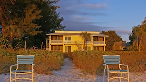 A beachfront property with a pathway leading to a building with a balcony. There are two chairs facing the building.