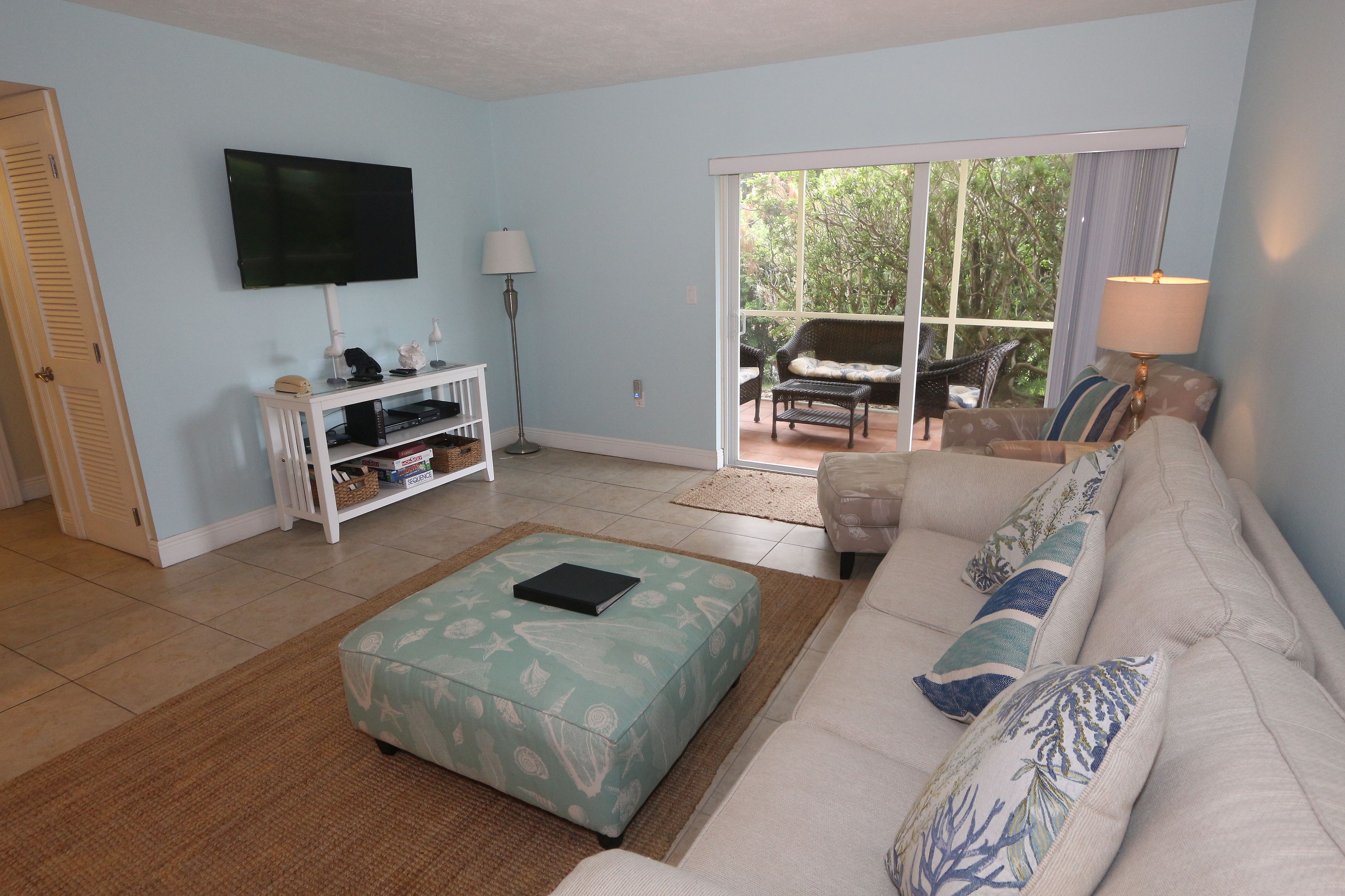A living room with a sofa, ottoman, flat-screen TV, and a window with a view of greenery.