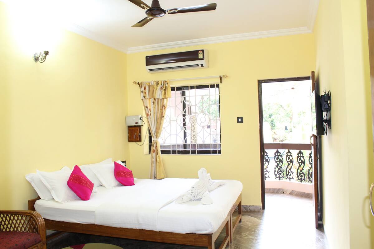 Habitación de hotel con cama, ventilador de techo, unidad de aire acondicionado, ventana con cortinas y puerta que da al exterior.
