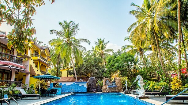Una piscina con sillas de descanso y sombrillas, rodeada de palmeras y un edificio con balcones.