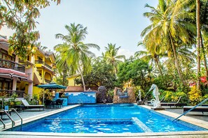 A swimming pool with lounge chairs and umbrellas, surrounded by palm trees and a building with balconies. - Sea View Resort (Canacona)
