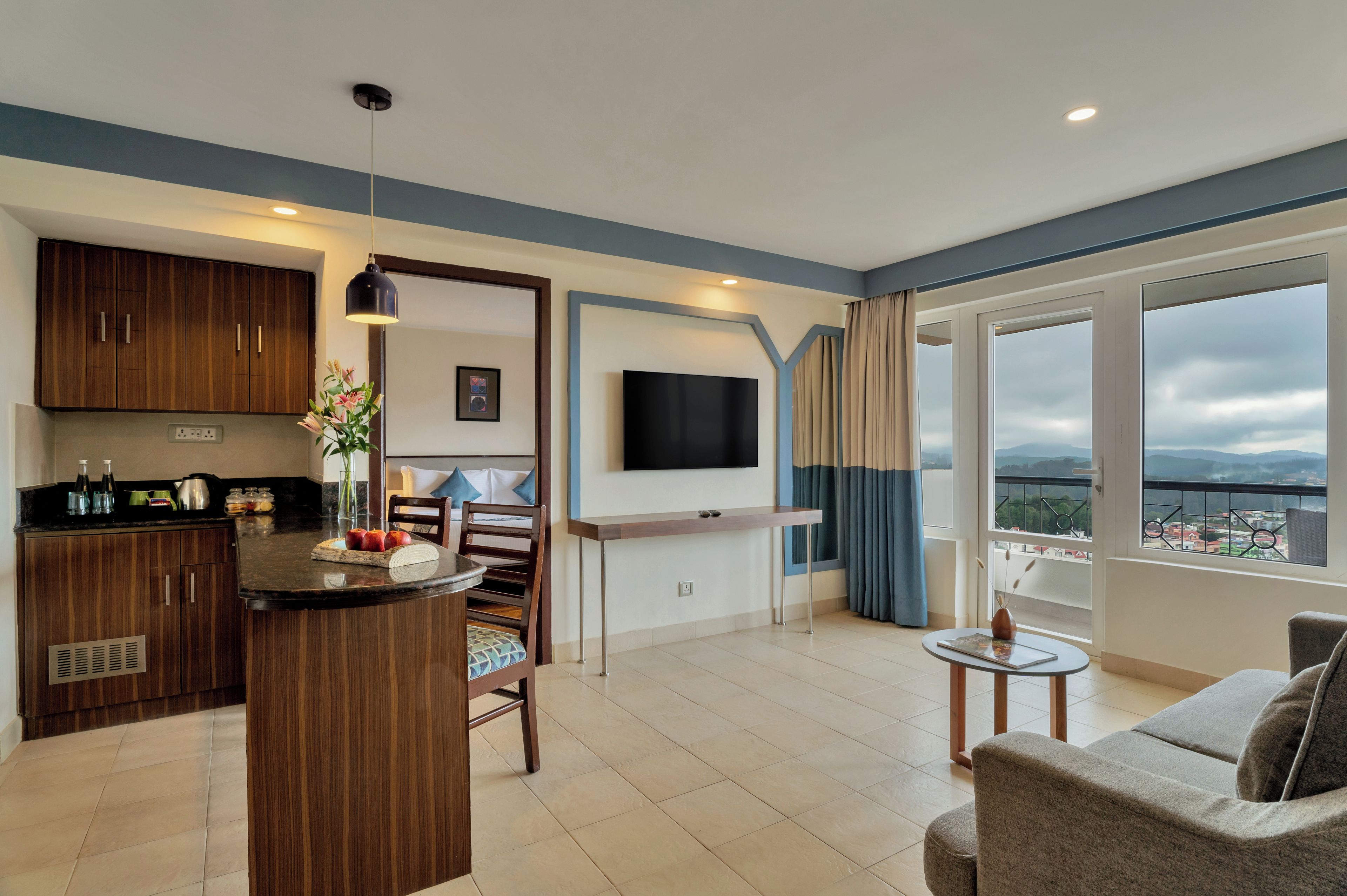 A modern living room with a kitchen area, a flat-screen TV mounted on the wall, and a balcony with a view.