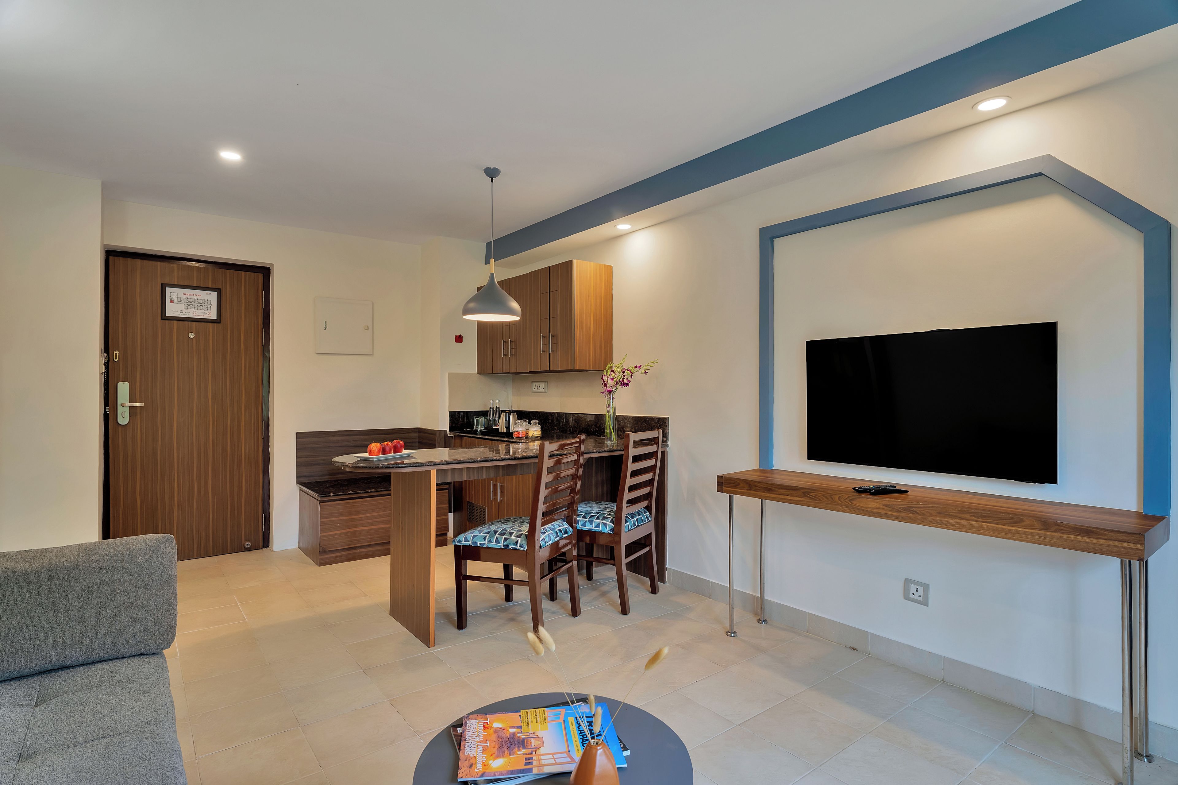 A modern living room with a flat-screen TV, a dining area with wooden chairs, and a kitchen with a wooden cabinet.