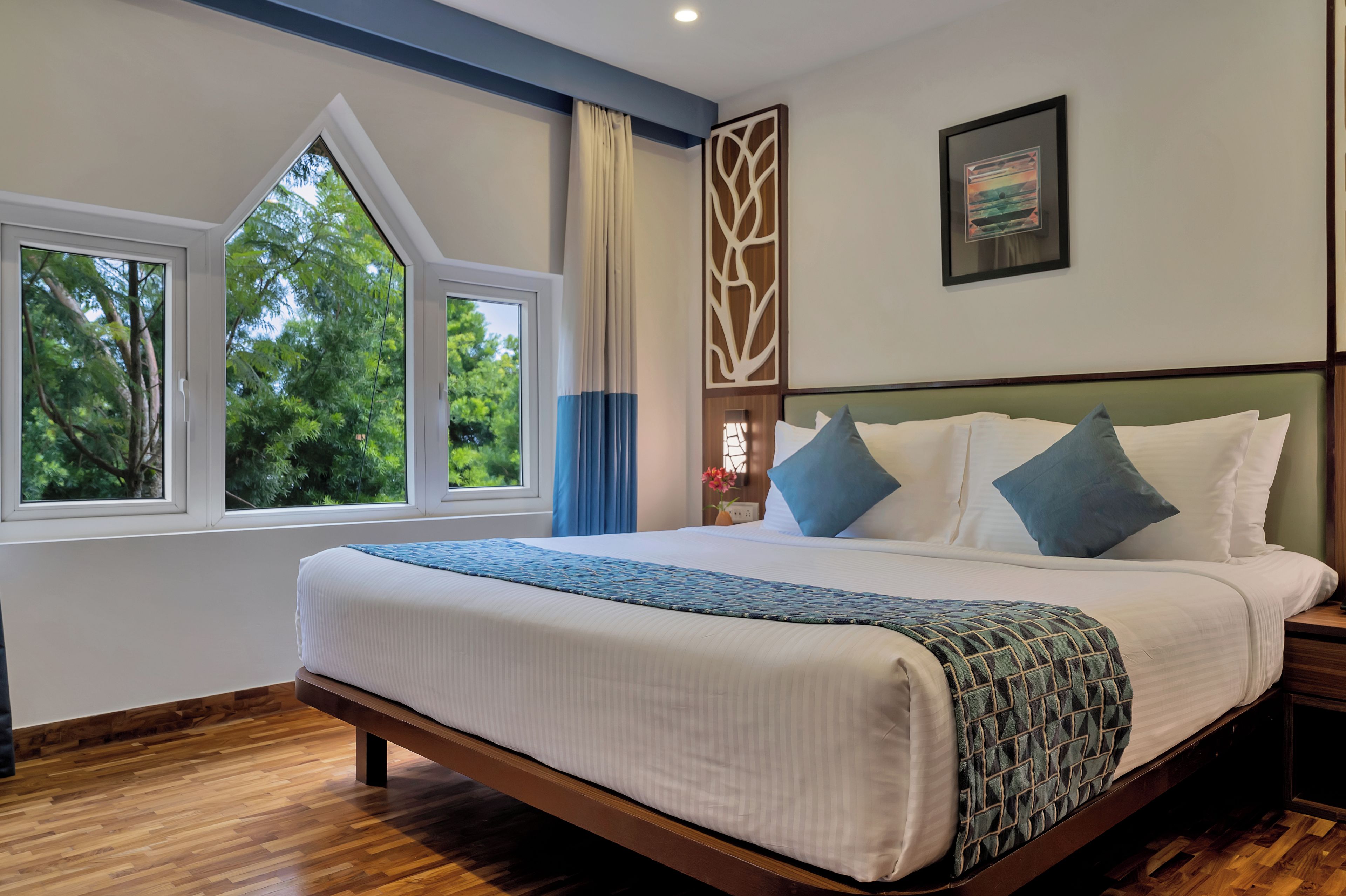 Habitación de hotel con una cama grande, dos ventanas, suelo de madera y una obra de arte colgada en la pared.