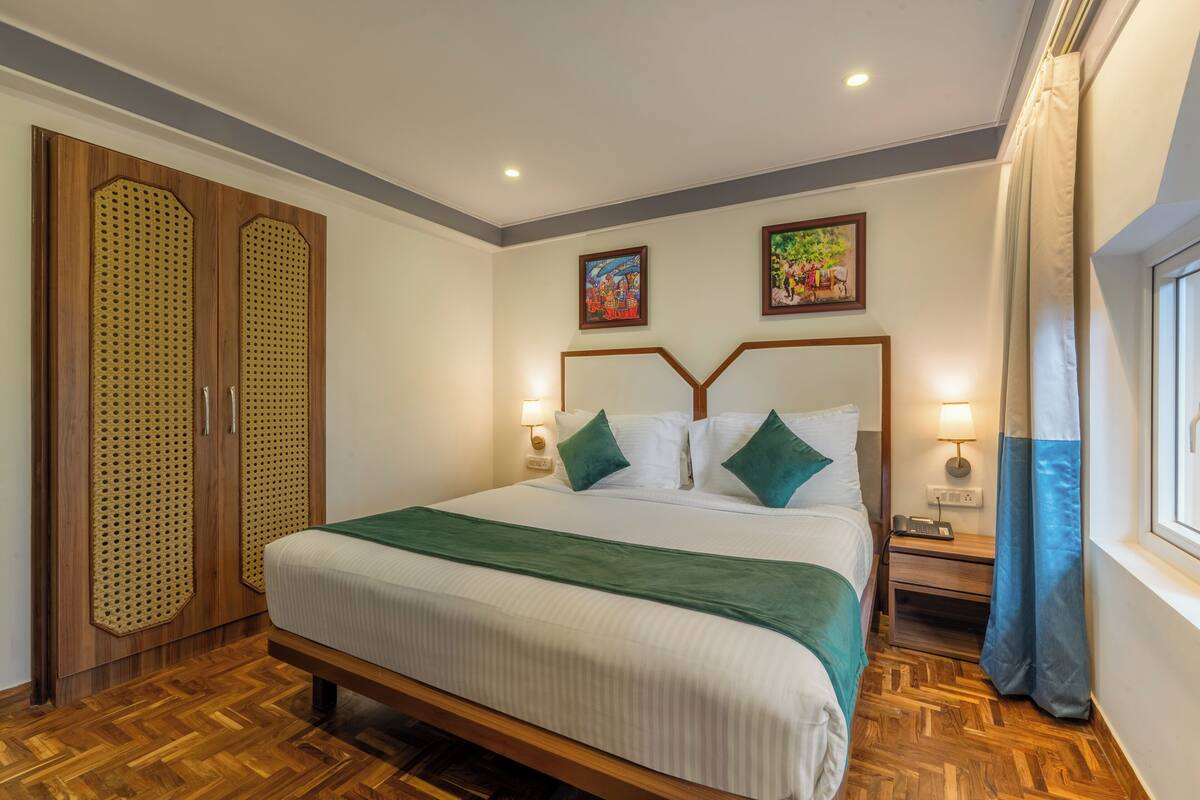 Une chambre d’hôtel bien aménagée, avec un grand lit, une armoire en bois, deux tableaux accrochés au mur, une table de chevet avec une lampe et une fenêtre avec des rideaux.