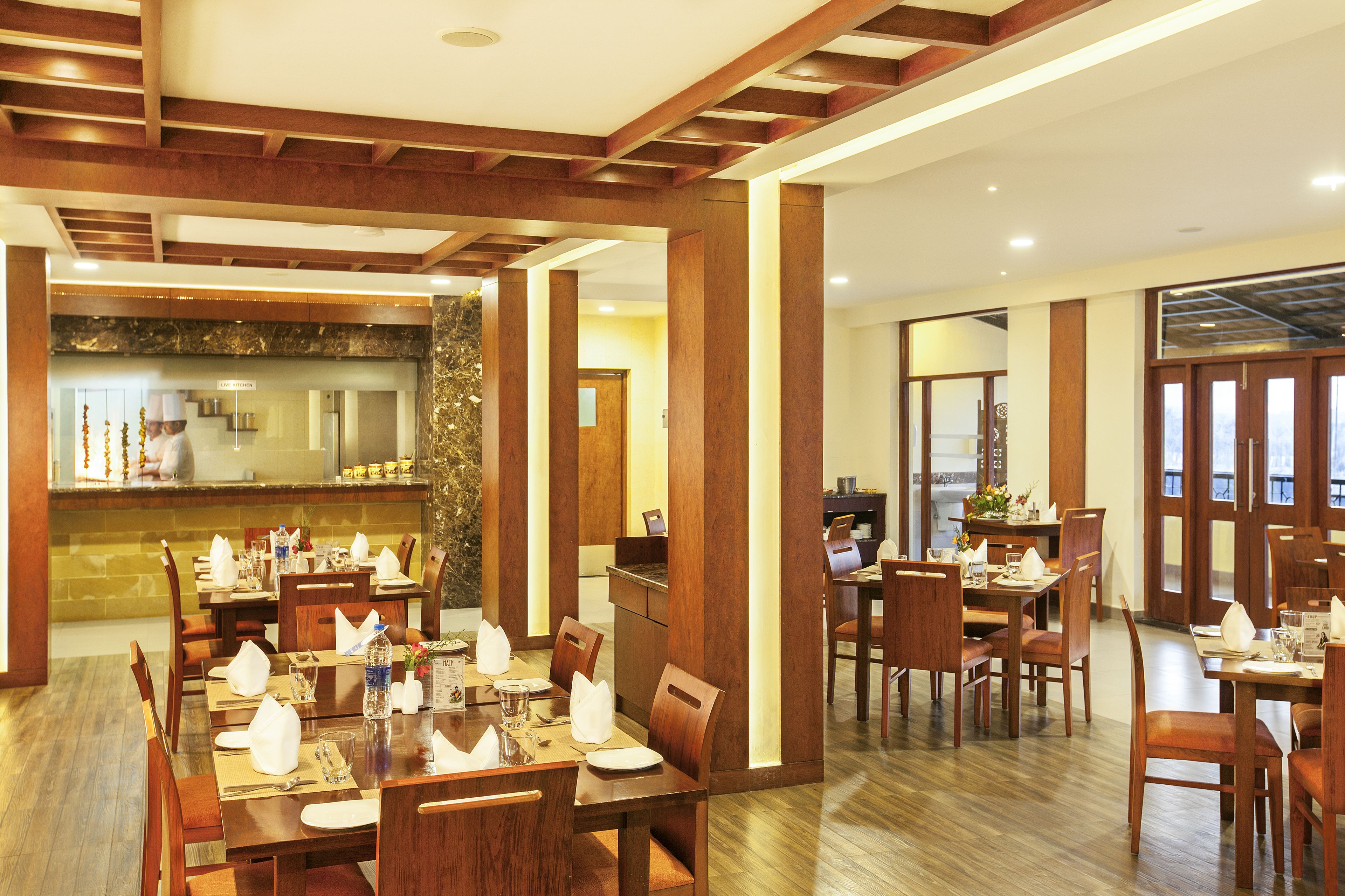A spacious dining area with wooden tables and chairs, a kitchen in the background, and a view of the outdoors through large windows.