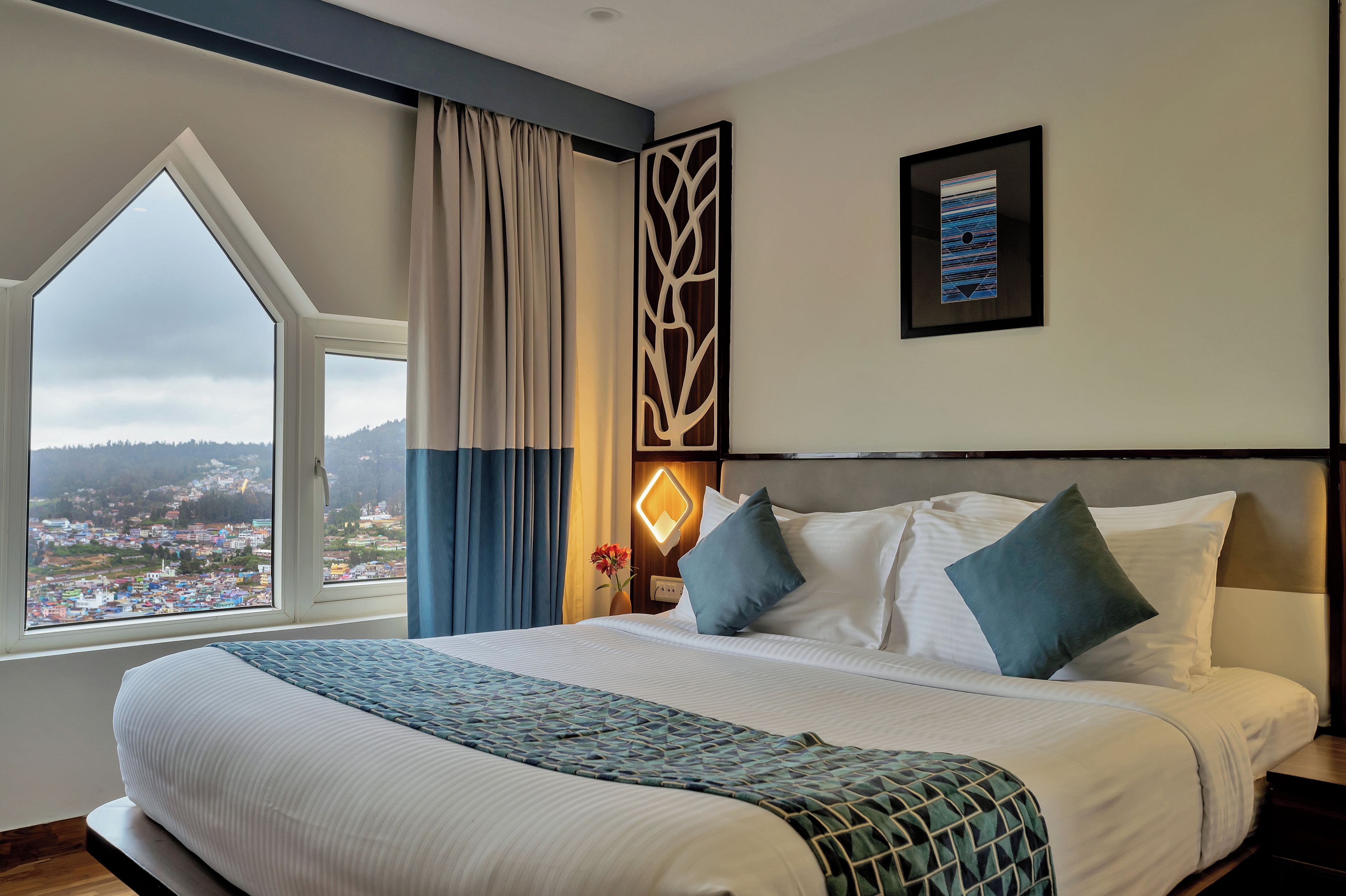 A hotel room with a large bed, a view of a cityscape through a window, and a decorative wall panel.