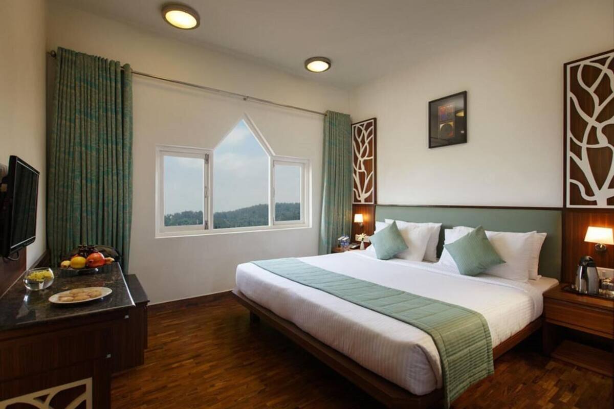 Une chambre d’hôtel avec un grand lit, un bureau agrémenté d’un panier de fruits, une fenêtre donnant sur un beau paysage et un élément décoratif mural.