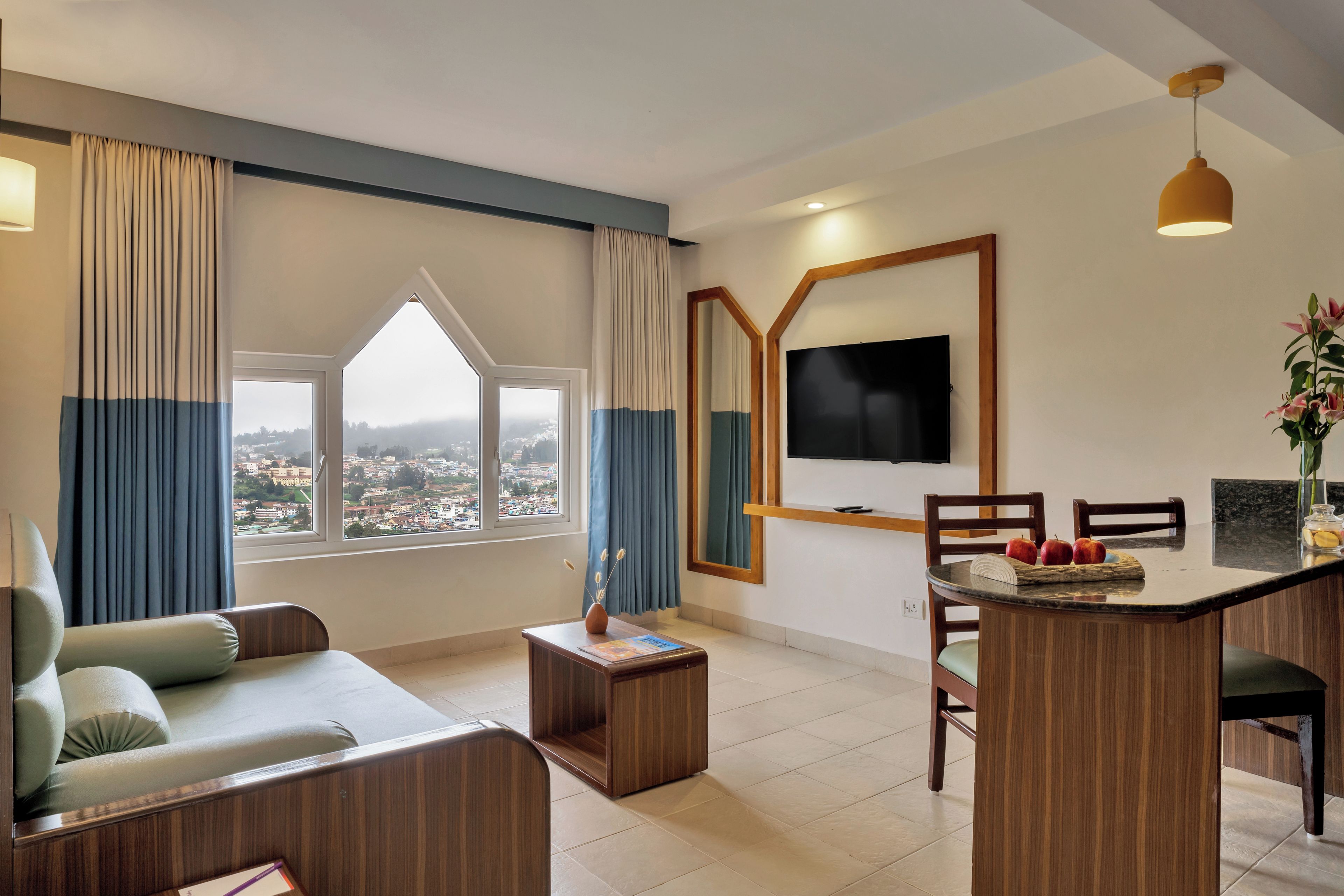 A modern living room with a sofa, a coffee table, a dining area with chairs, a TV mounted on the wall, and a view of the city through large windows.