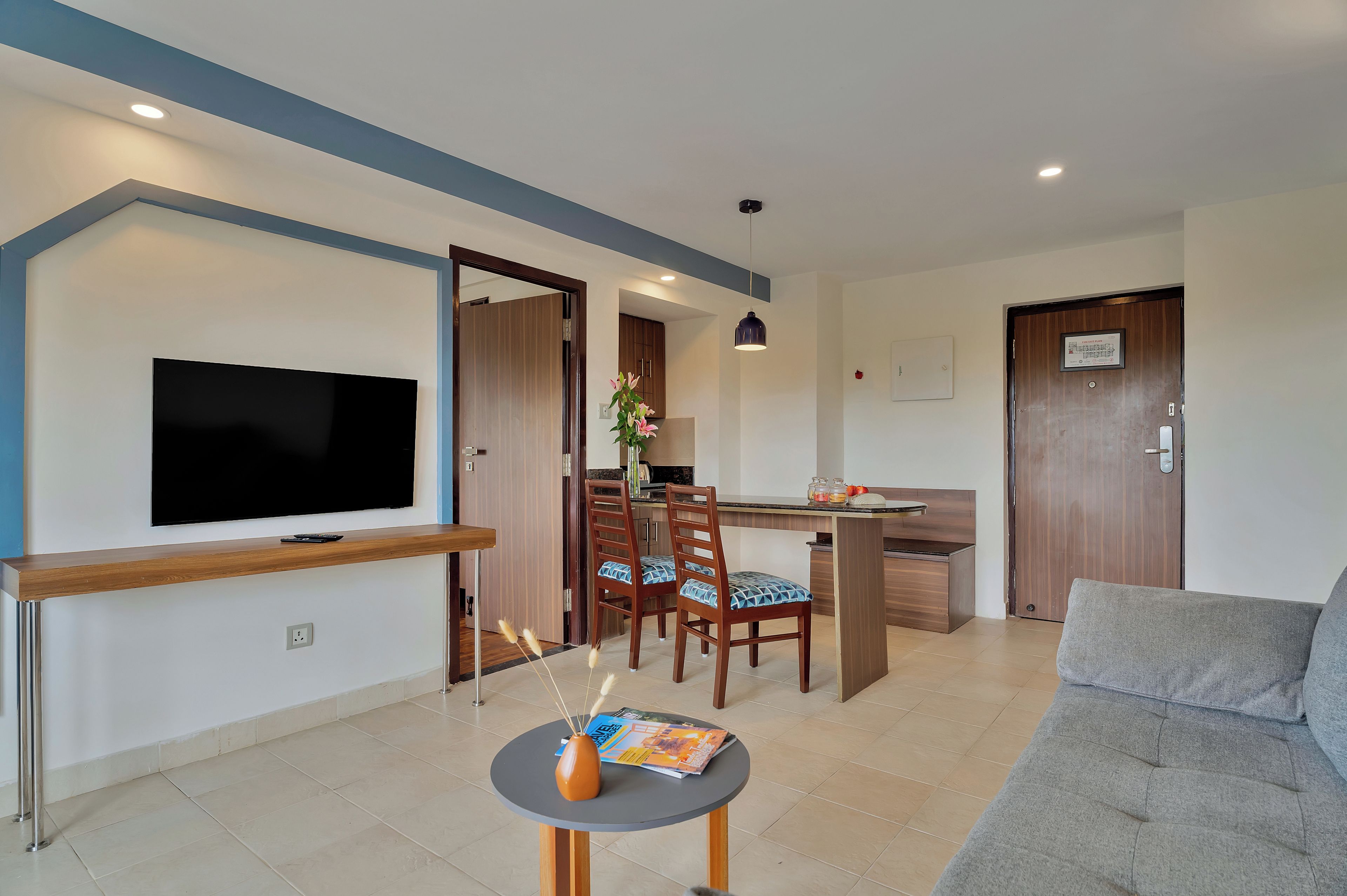 A modern living room with a flat-screen TV, a grey sofa, a dining area with a wooden table, and a kitchen visible through an open door.