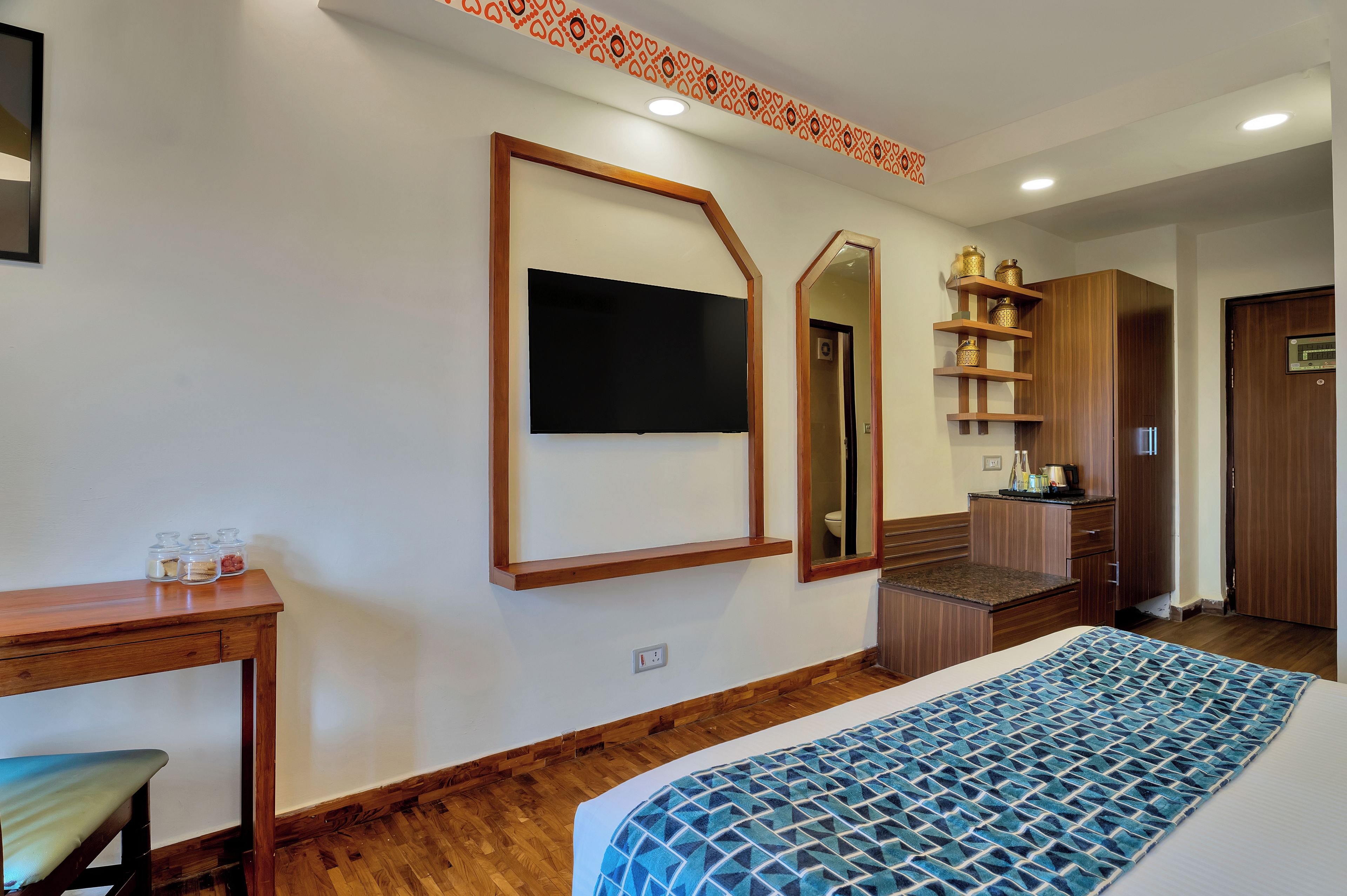 A hotel room with a bed, a desk, a television, a mirror, a shelf with decorative items, and a wardrobe.