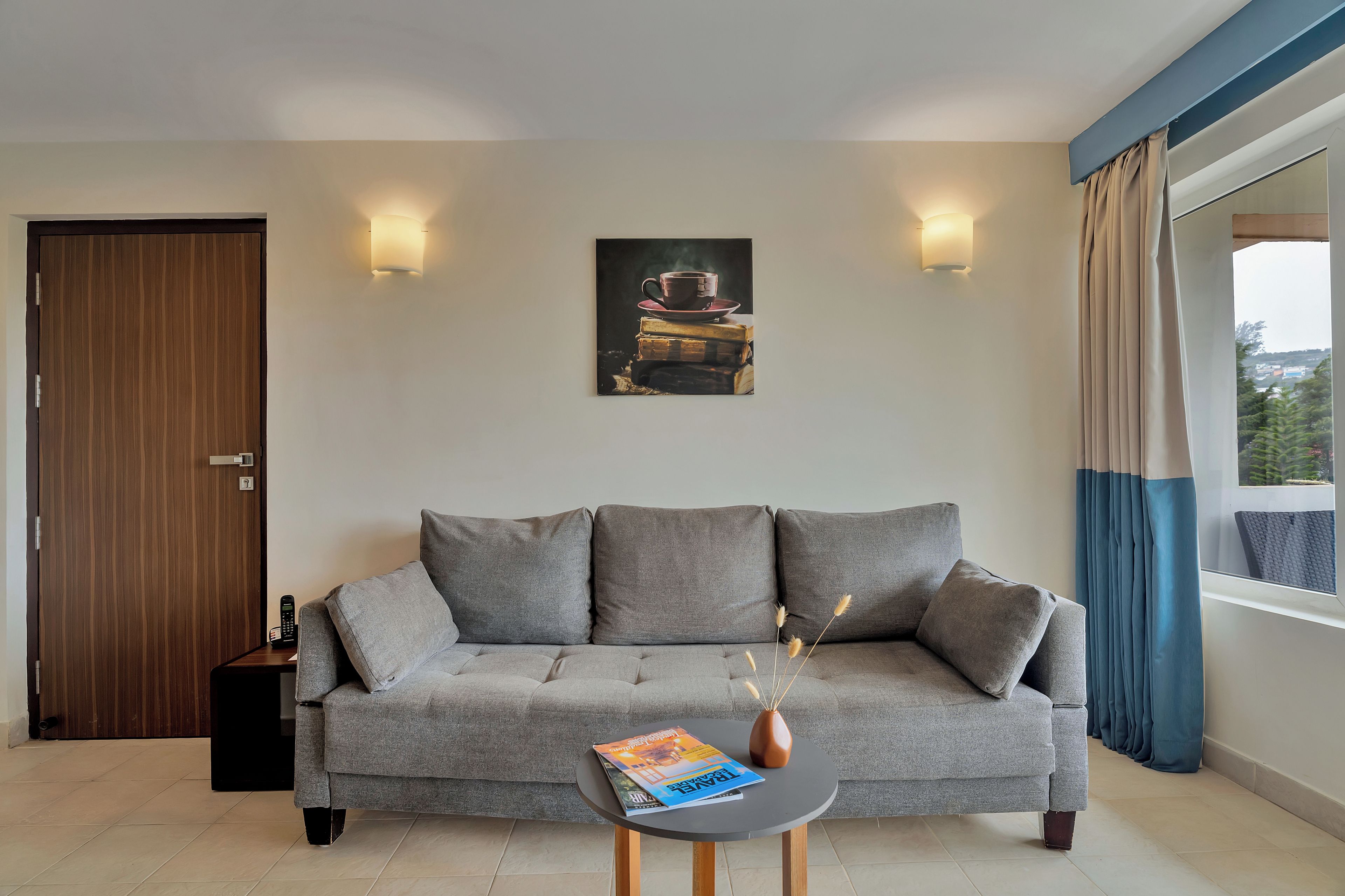 Una sala de estar moderna con un sofá gris, una mesa de centro con revistas, un cuadro sobre el sofá y vistas al exterior por la ventana.