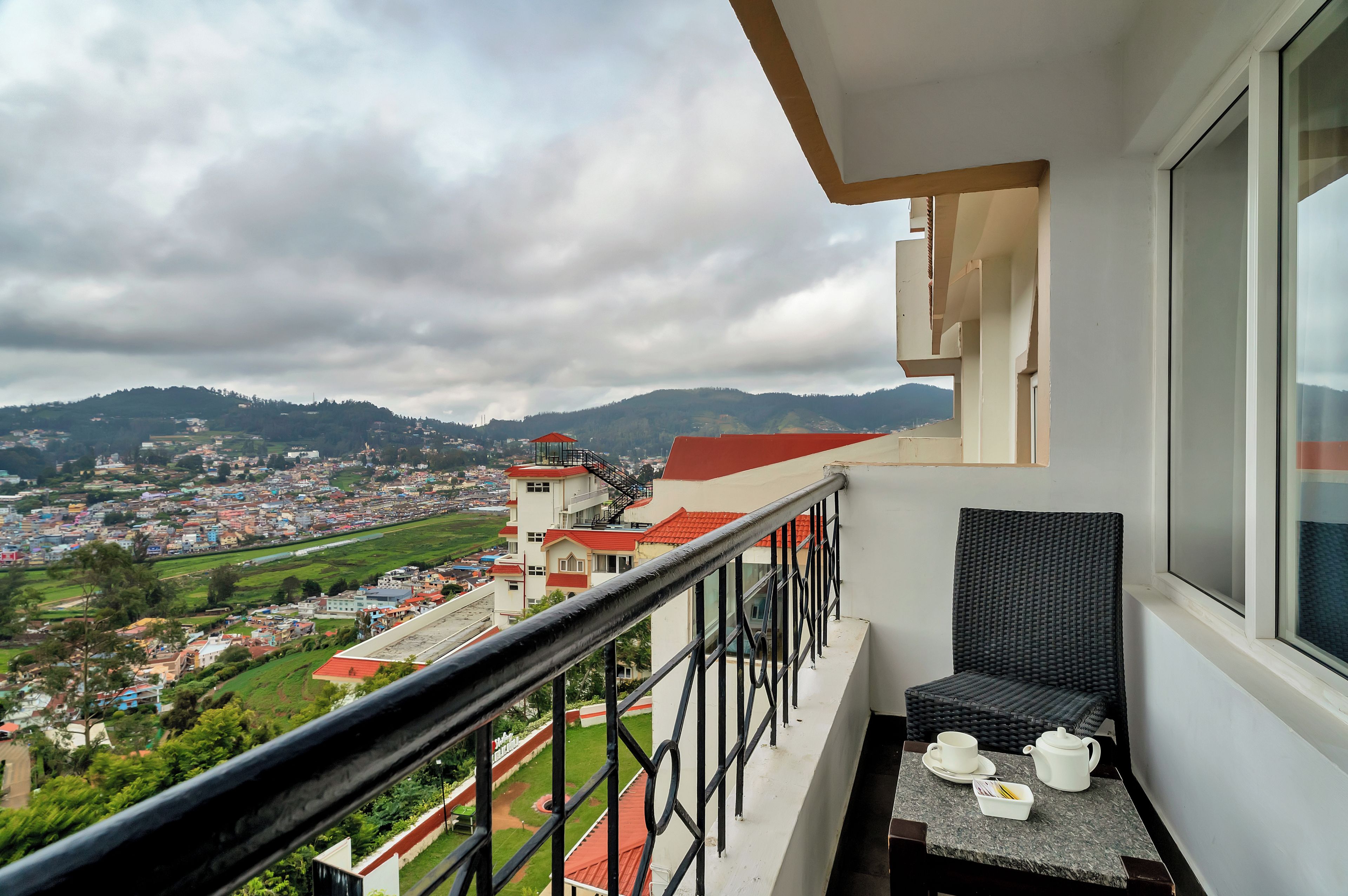 A balcony with a view of a town and hills, a table with a teapot and cups, and a wicker chair.
