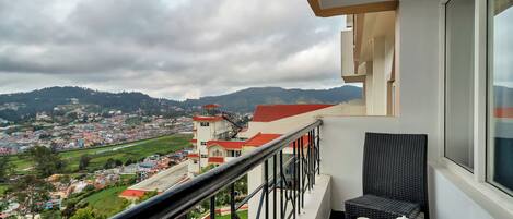 A balcony with a view of a town and hills, a table with a teapot and cups, and a wicker chair.