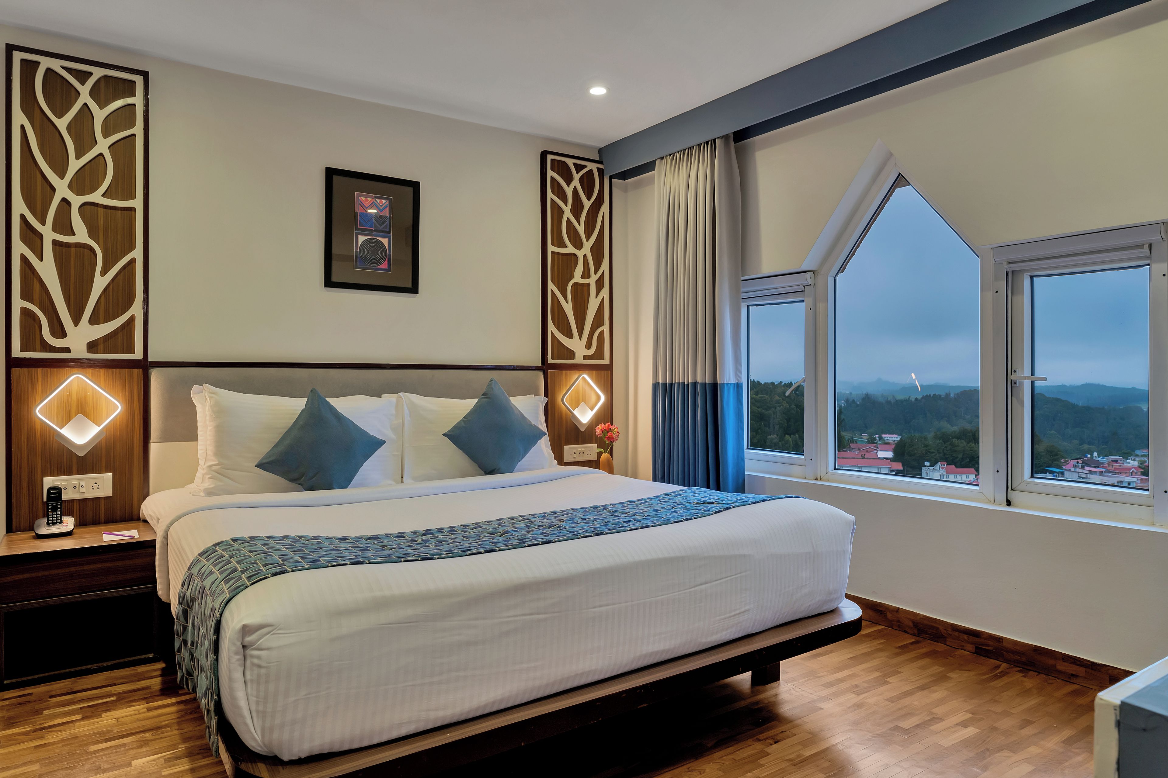 A hotel room with a large bed, a bedside table with a phone, a window with a view of the landscape, and decorative wall panels.