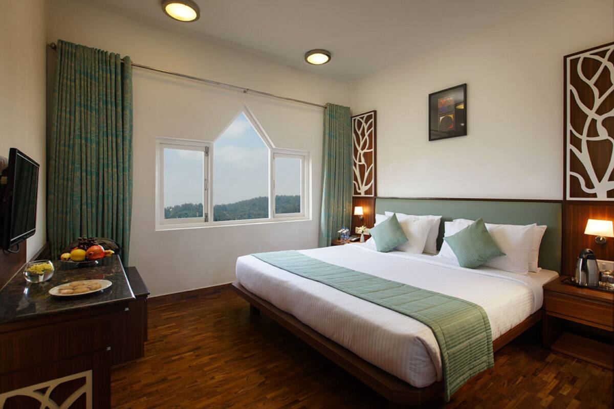 Une chambre d’hôtel avec un grand lit, un bureau agrémenté d’un panier de fruits, une fenêtre avec des rideaux et une vue sur l’extérieur.