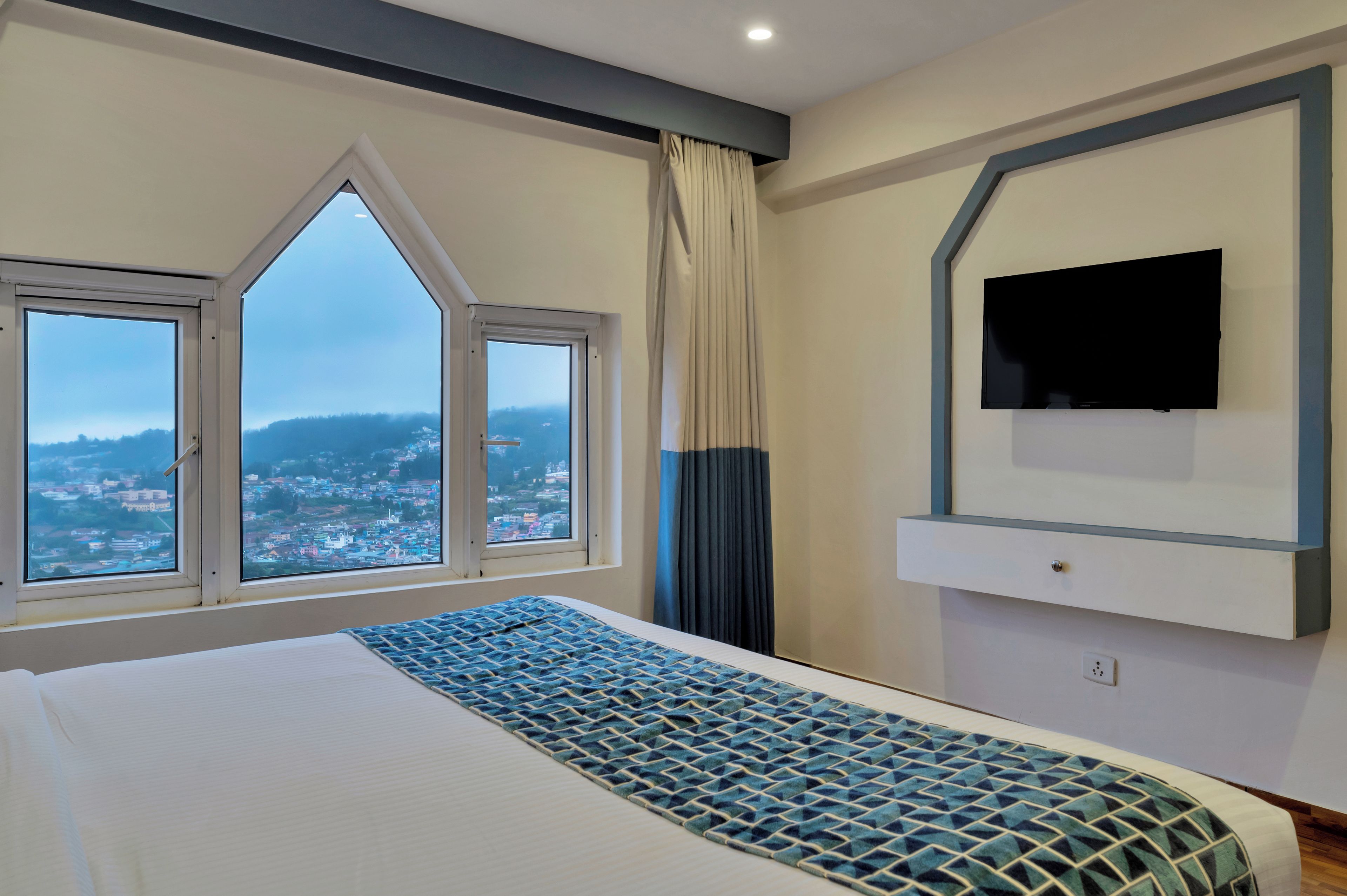 A hotel room with a large bed, a flat-screen TV mounted on the wall, and a view of a cityscape through a set of windows.