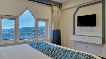 A hotel room with a large bed, a flat-screen TV mounted on the wall, and a view of a cityscape through a set of windows.