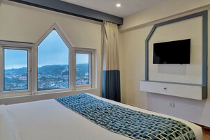 A hotel room with a large bed, a flat-screen TV mounted on the wall, and a view of a cityscape through a set of windows.