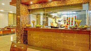 Un mostrador de recepción de mármol con un separador de vidrio, una cocina al fondo, y diversas frutas y verduras expuestas en la encimera.