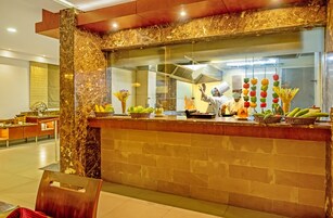 Un mostrador de recepción de mármol con un separador de vidrio, una cocina al fondo, y diversas frutas y verduras expuestas en la encimera.