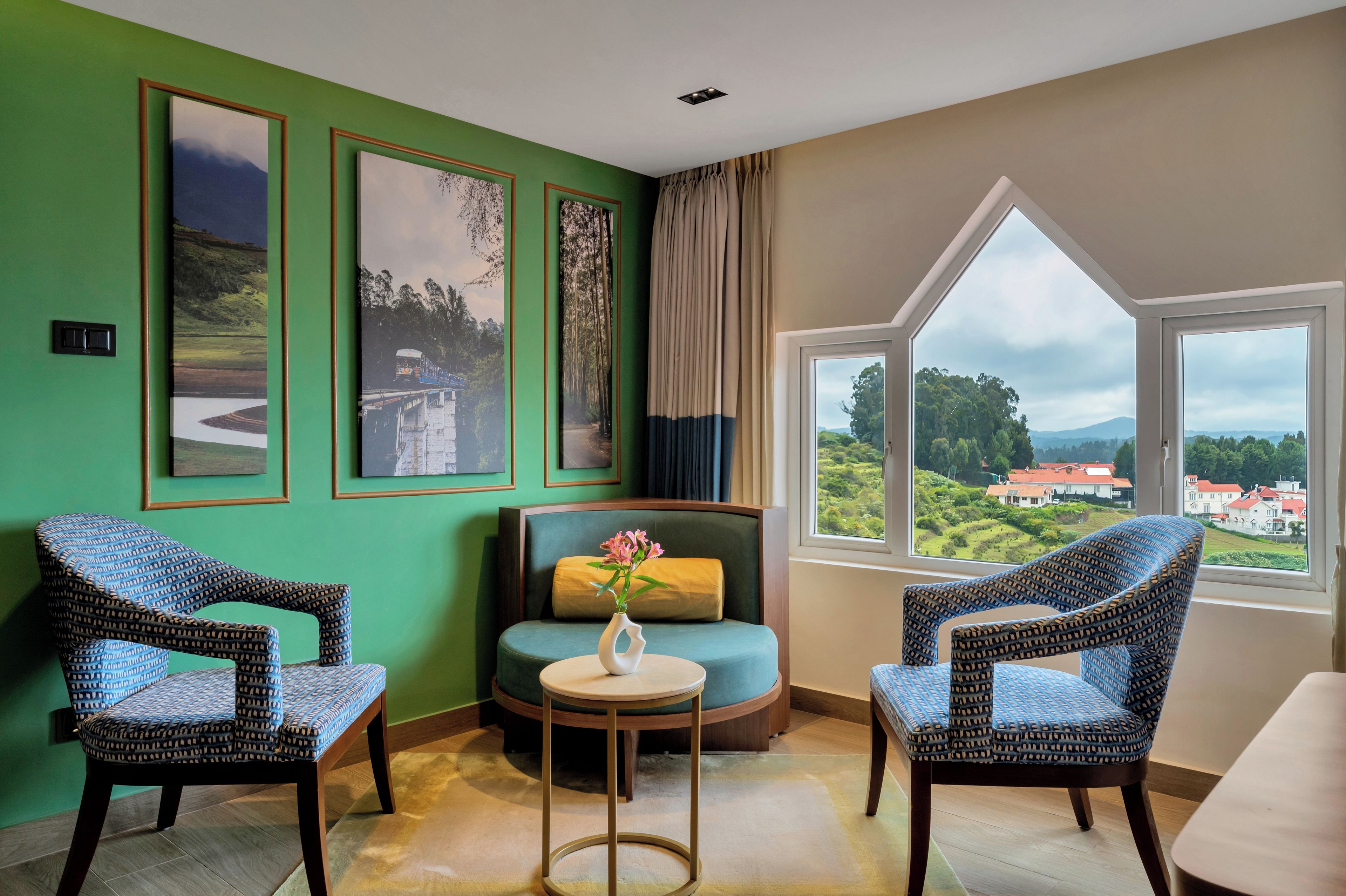 Une chambre d’hôtel avec un mur vert, deux fauteuils à motifs, une petite table ronde avec un vase, un canapé avec un coussin et une grande fenêtre donnant sur un paysage verdoyant et des bâtiments.