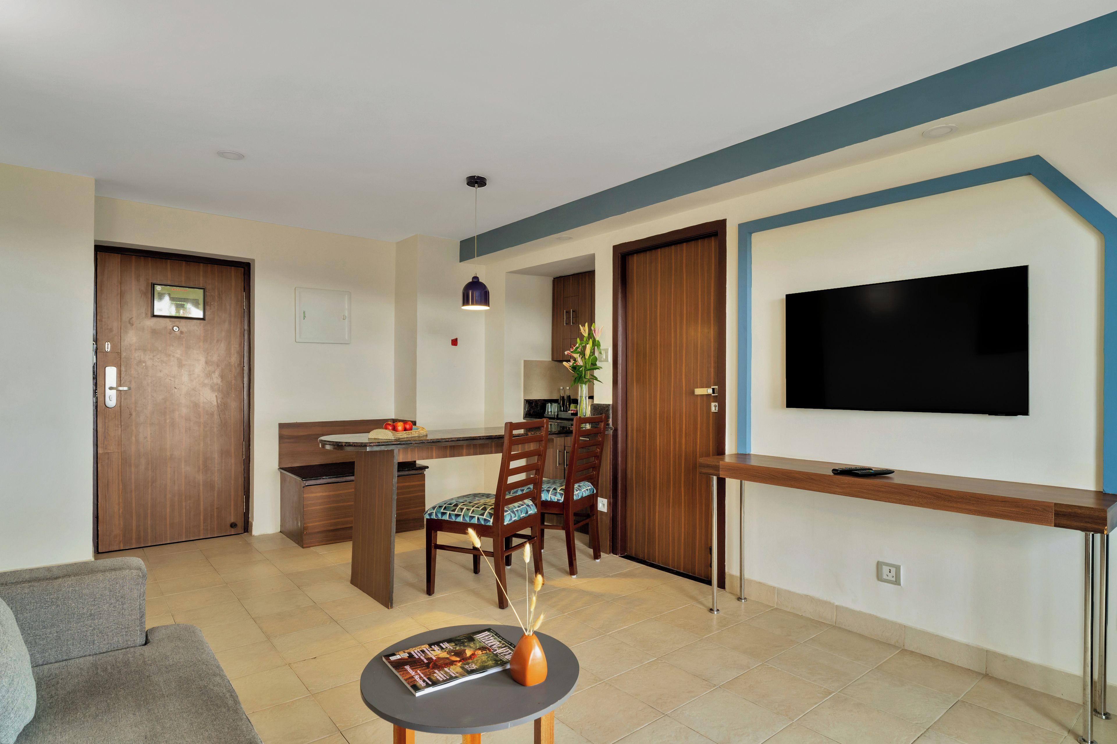 A modern living room with a flat-screen TV, a dining area with chairs and a table, and a kitchen area with cabinets and a countertop.