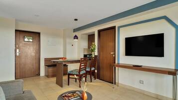 A modern living room with a flat-screen TV, a dining area with chairs and a table, and a kitchen area with cabinets and a countertop.