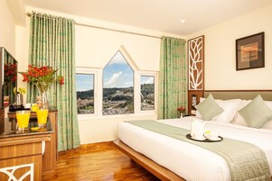 A hotel room with a large bed, a view of the city through a window, a wooden desk with a vase of flowers, and a tray with breakfast items.