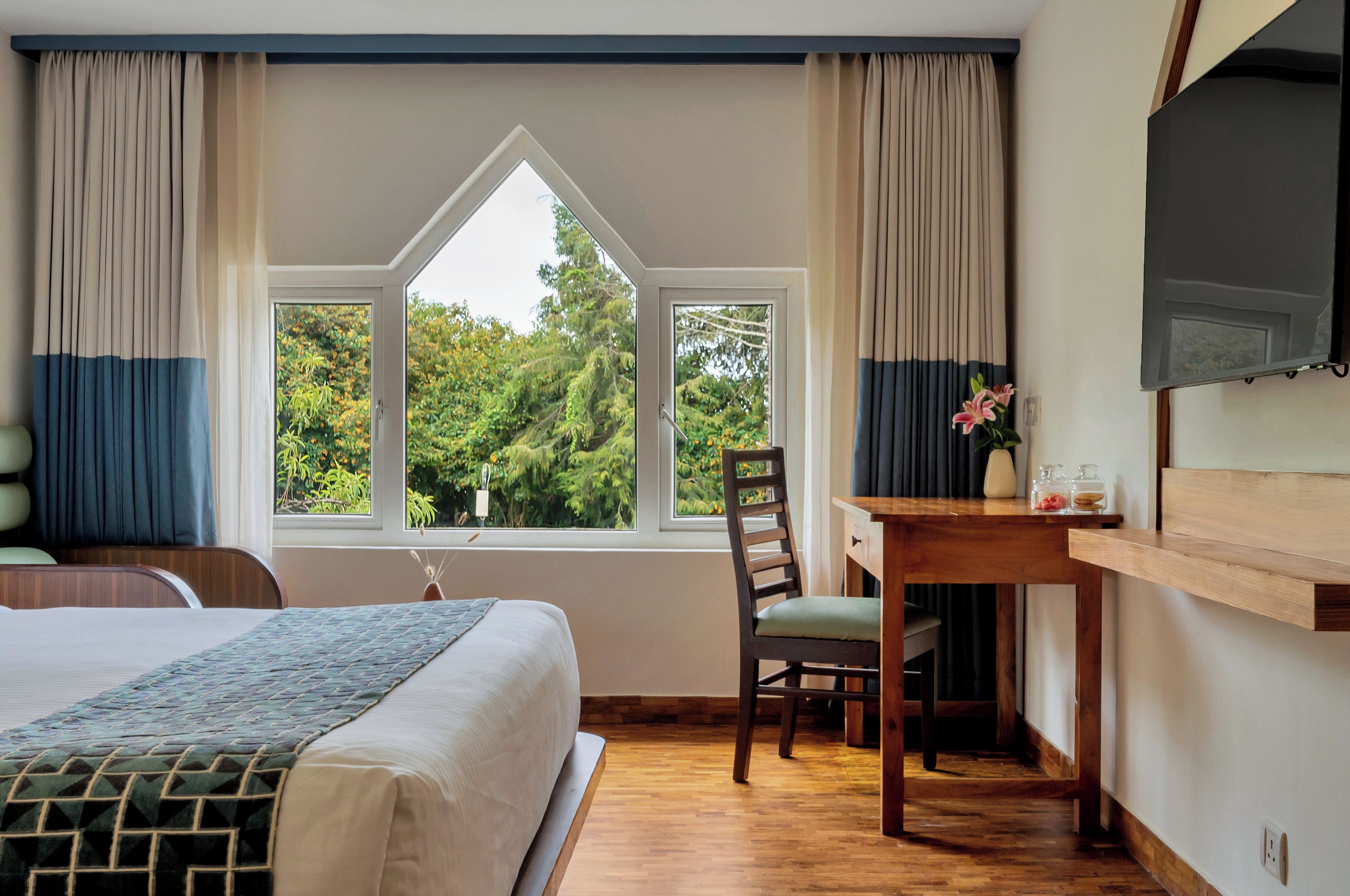 A bedroom with a bed, a desk, and a chair. There is a window with curtains and a view of trees.
