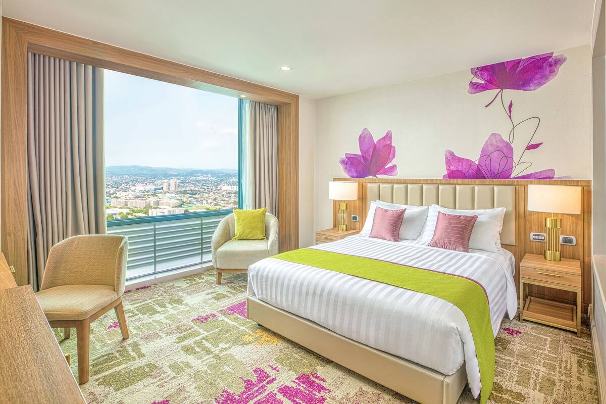 Hotelkamer met een groot bed, twee nachtkastjes, een stoel en uitzicht op een stadsgezicht door grote ramen.