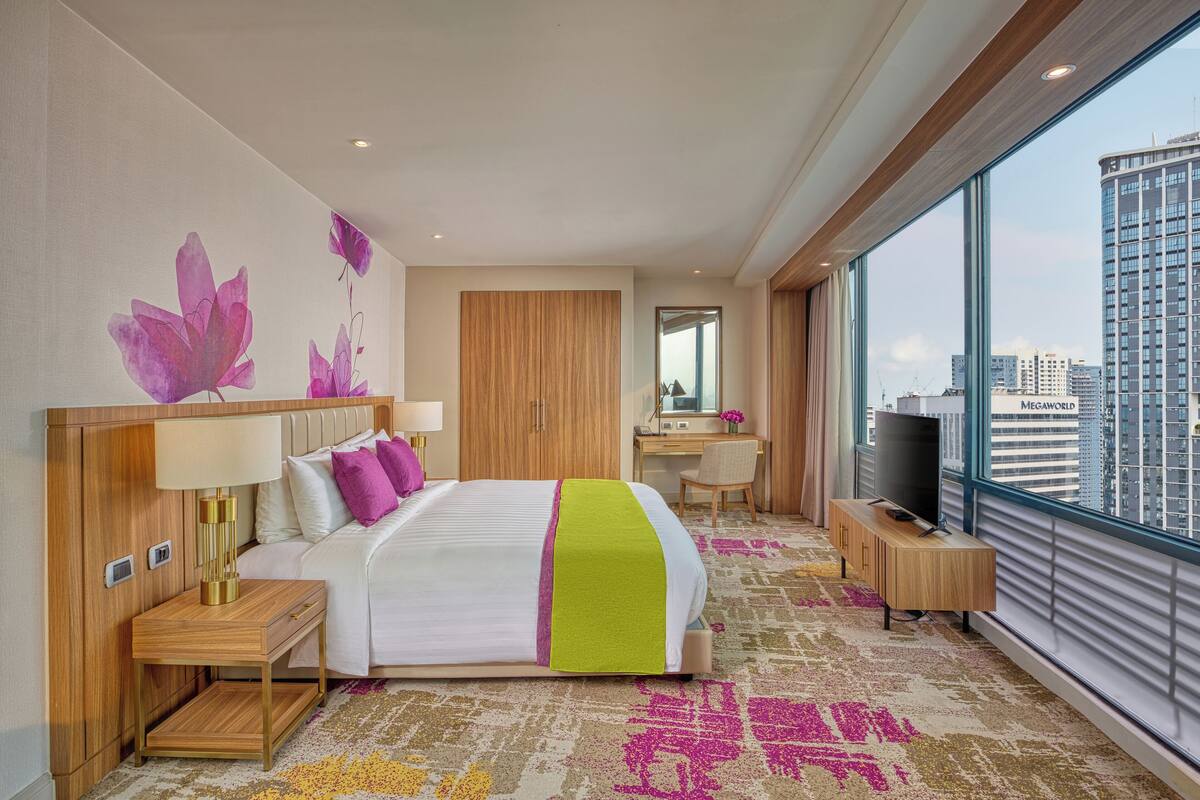 Een moderne hotelkamer met een groot bed, nachtkastjes, een televisie, een bureau en een groot raam met uitzicht op de stad.