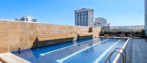 A rooftop swimming pool with clear blue water, a metal ladder, and a view of surrounding buildings.