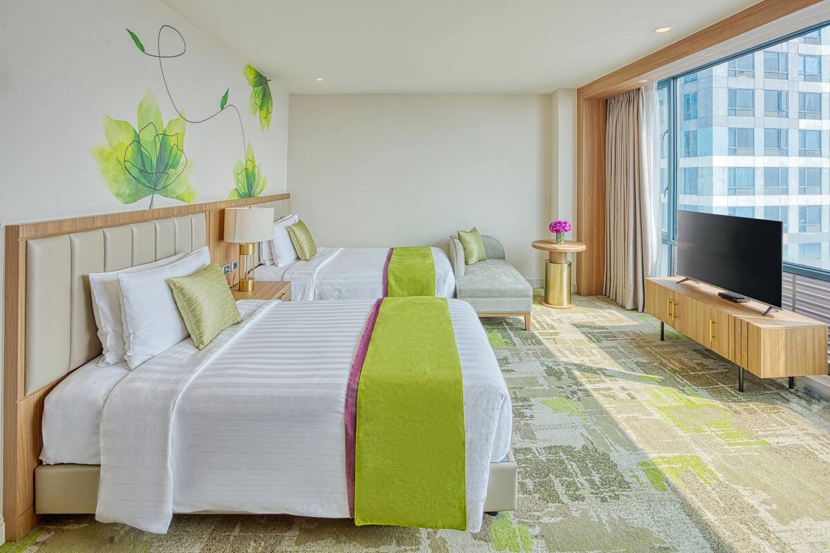 Hotelkamer met twee bedden, een televisie, een kleine tafel met een bloemenvaas en een groot raam met uitzicht over de stad.