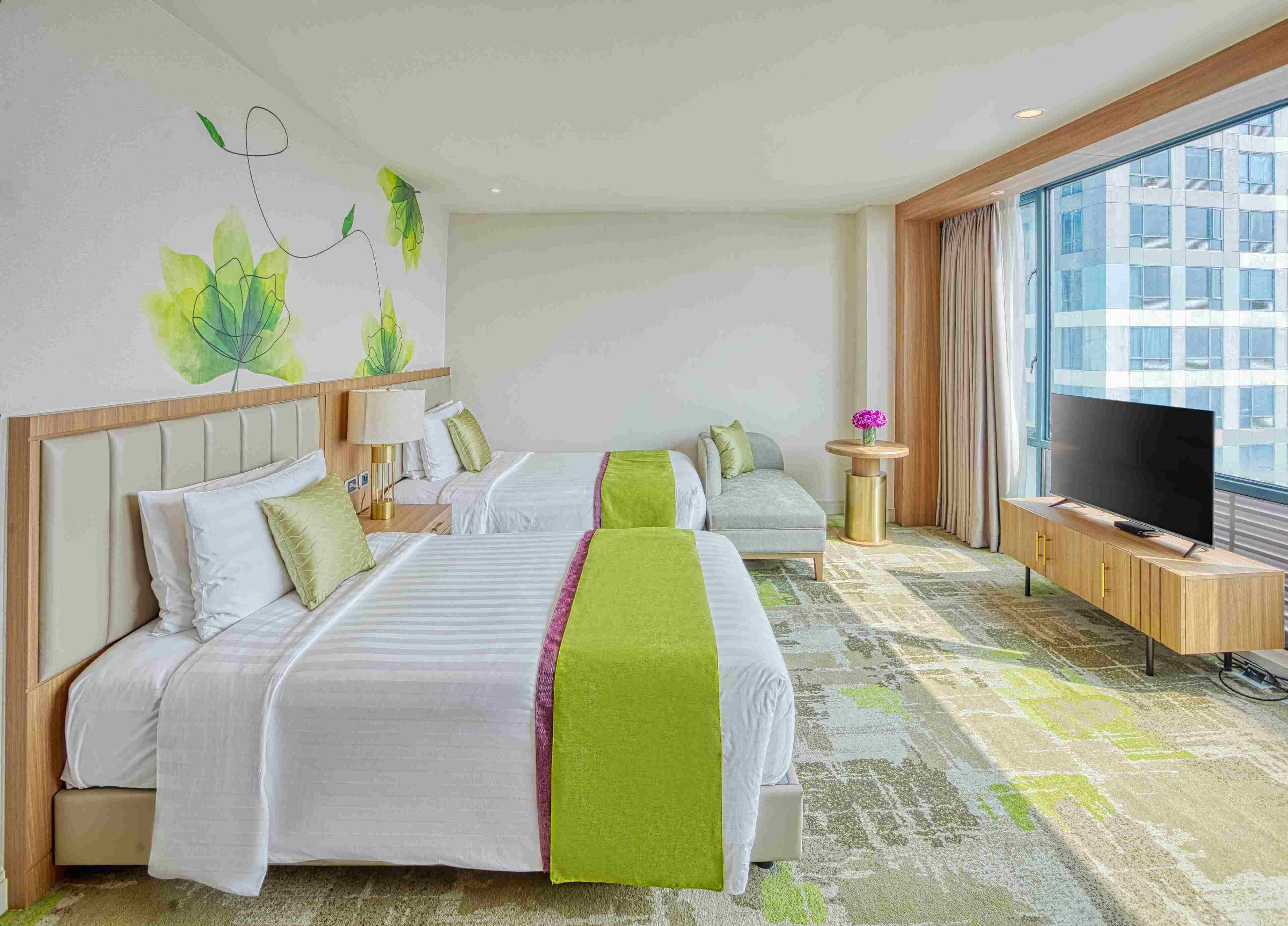 Hotellihuone, jossa on kaksi sänkyä, televisio, pieni pöytä maljakolla ja suuri ikkuna.