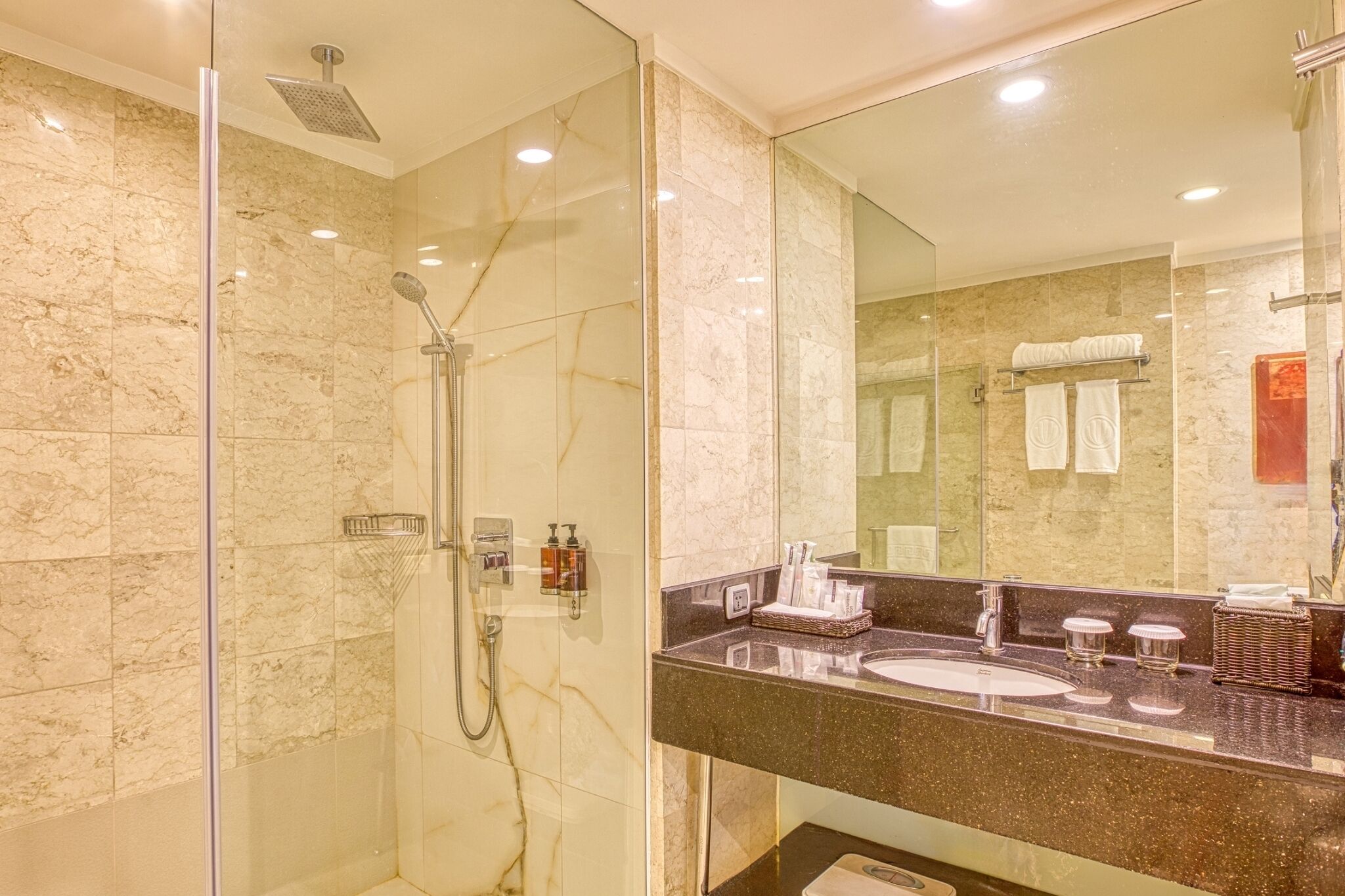 Een moderne badkamer met een douche met glazen wand, marmeren wanden en een grote spiegel boven een granieten aanrecht met een wastafel en handdoekhouder.