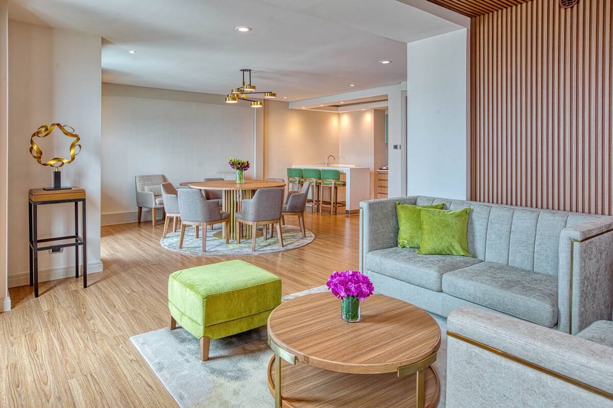 Een moderne woonkamer met een grijze bank, een groene poef en een ronde salontafel. Er is een eethoek met een ronde tafel en stoelen, en een keuken op de achtergrond.