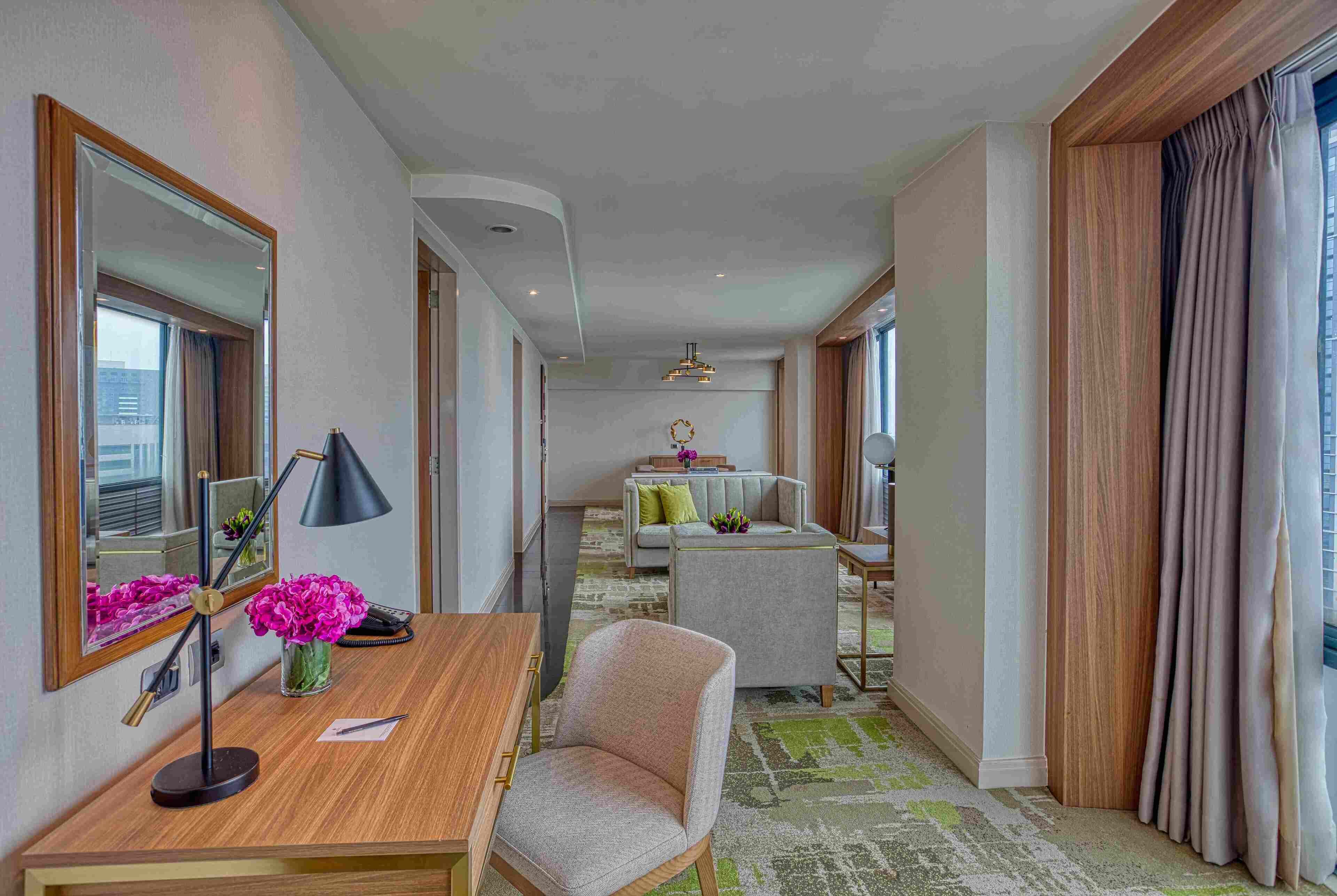 Habitación de hotel con escritorio de madera, una silla, un sofá, un espejo grande y vistas a la ciudad.