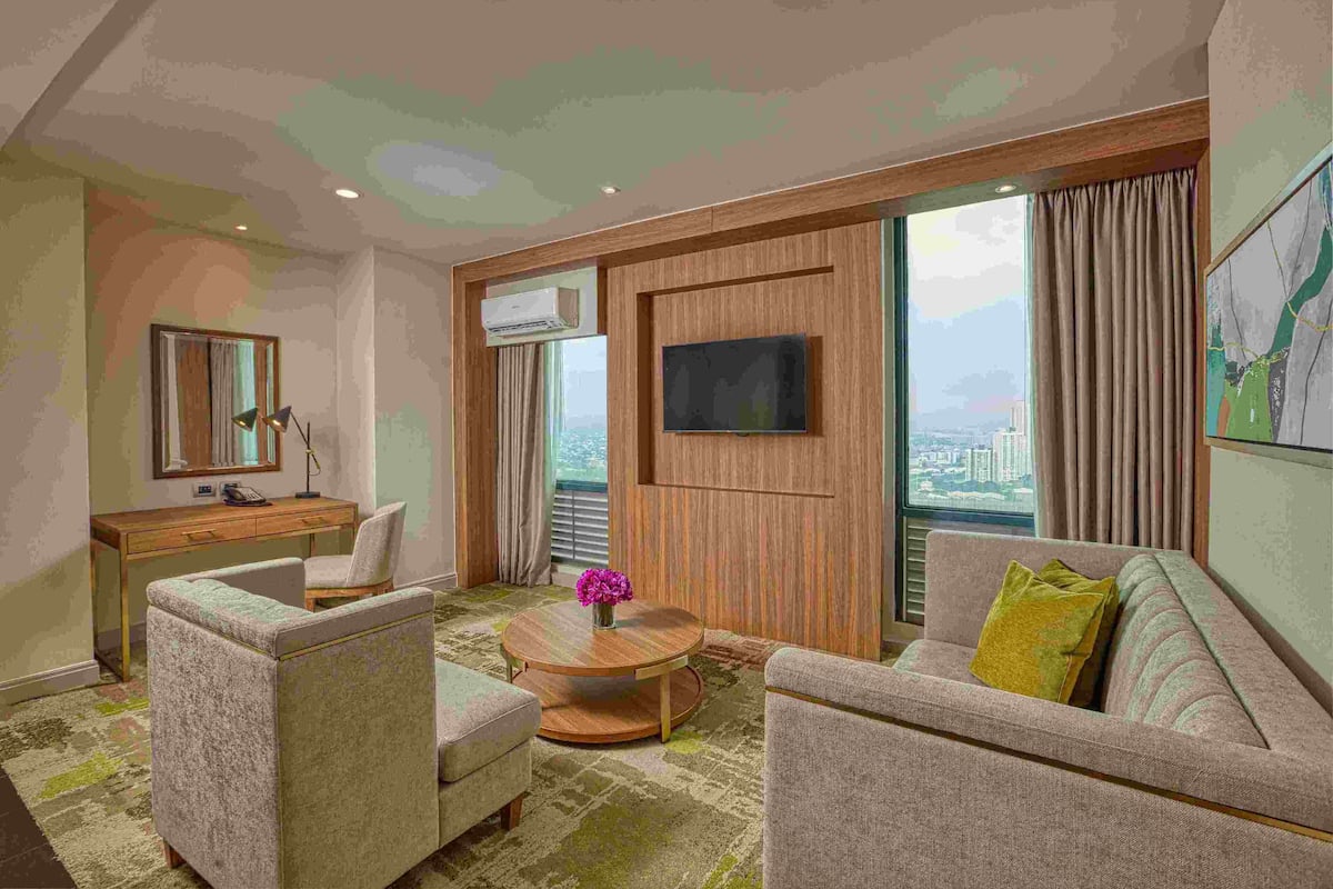 Een hotelkamer met een houten bureau, twee fauteuils, een ronde salontafel met een bloevas, een flatscreen televisie aan de muur en grote ramen met uitzicht op de stad.