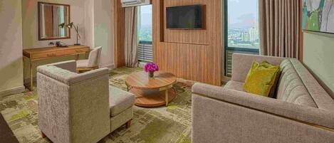 Une chambre d’hôtel dotée d’un bureau en bois, de deux fauteuils, d’une table basse ronde agrémentée d’un vase de fleurs, d’un téléviseur à écran plat fixé au mur et de grandes fenêtres offrant une vue sur la ville.