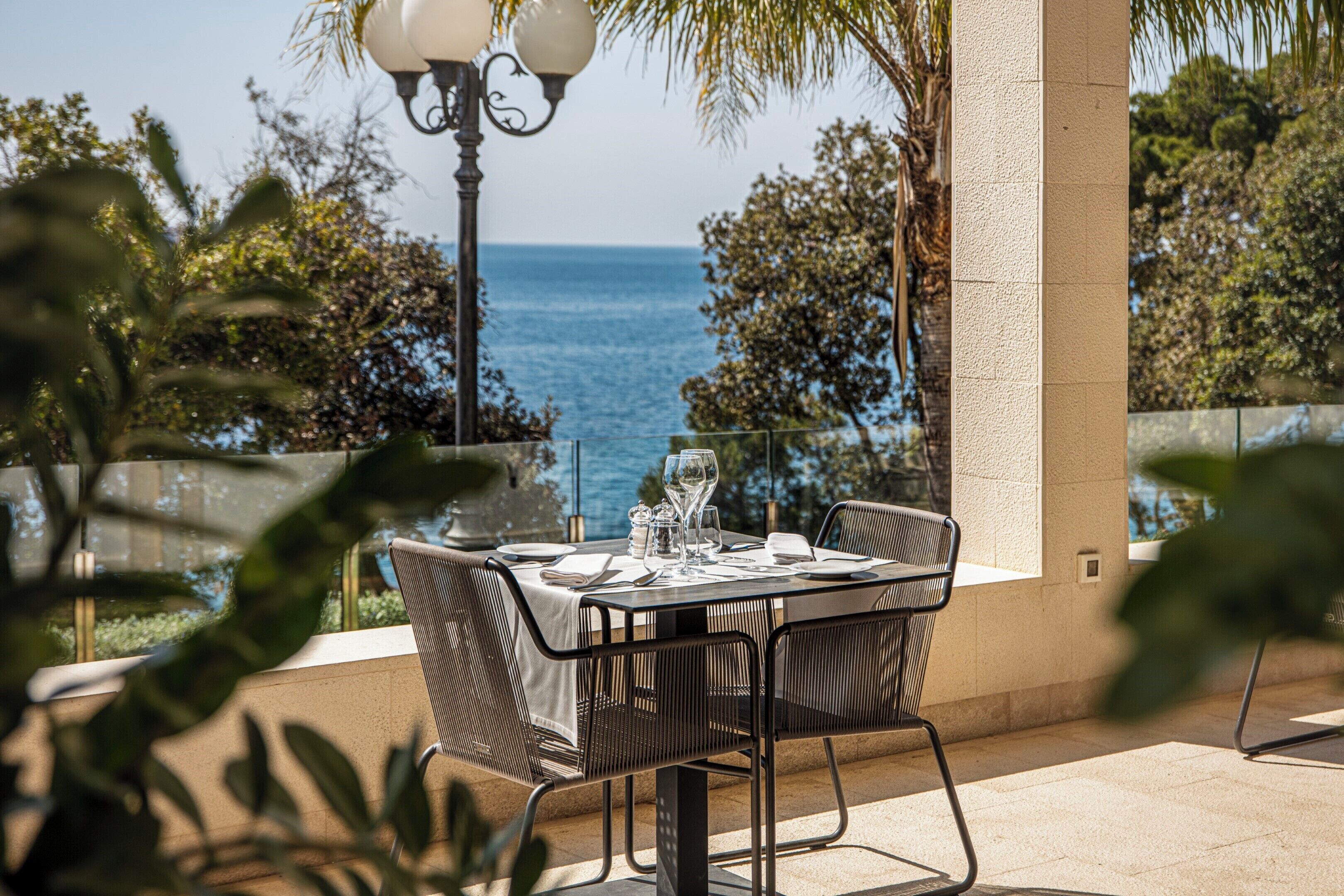 A table set for dining with a view of the sea, surrounded by greenery and a lamp post.